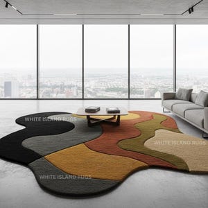 May include: An abstract area rug with a unique, organic shape. The rug features a color palette of black, gray, yellow, orange, and olive green. The rug is in a modern living room setting with large windows and a gray sofa. The text "WHITE ISLAND RUGS" is visible.