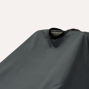 May include: A dark gray barber cape with a black neckband and a black strap. The cape is made of a smooth, water-resistant material. The cape is designed to protect clothing during haircuts and styling.