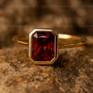 Könnte beinhalten: Ein goldener Ring mit einem rechteckigen, tiefroten Edelstein. Der Edelstein ist in einer goldenen Lünette gefasst, und das Band ist aus poliertem Gold. Der Ring wird auf einer strukturierten, braunen Oberfläche präsentiert.