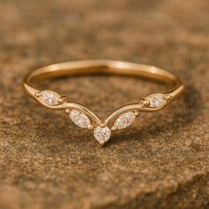 May include: A gold-colored ring with a curved design. The ring features five clear, sparkling gemstones. The gemstones are set in a delicate, elegant pattern. The ring is displayed on a textured, brown surface.