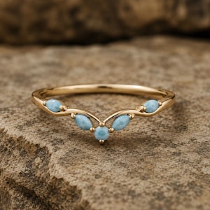 Peut inclure: Une bague dorée avec un design incurvé, ornée de cinq pierres précieuses bleu clair. La bague est posée sur une surface de pierre brune texturée, mettant en valeur les détails délicats et le savoir-faire de la bijouterie.