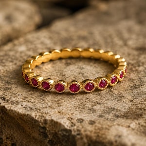 May include: A gold band ring featuring a continuous row of small, round, vibrant pinkish-red gemstones set in individual scalloped metal settings that encircle the entire circumference of the band, creating a textured and detailed jewellery piece.