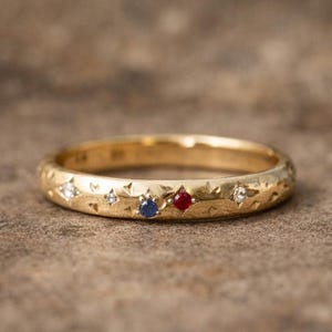 Op de afbeelding: Een gouden ring met een textuuroppervlak en ingelegde edelstenen. De ring heeft kleine witte, blauwe en rode edelstenen. De ring is geplaatst tegen een neutrale achtergrond.