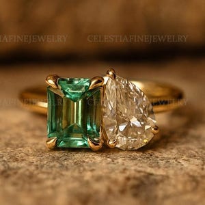 May include: A gold ring featuring an emerald-cut green gemstone and a pear-shaped clear gemstone. The ring is set on a textured, brown surface. The brand name "CELESTIAFINEJEWELRY" is visible in the background.