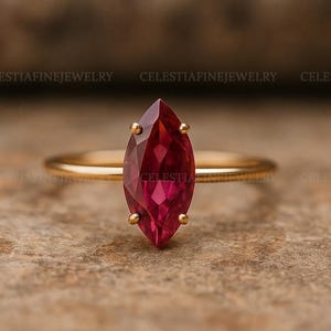 Op de afbeelding: Een gouden ring met een marquise-geslepen, dieprode edelsteen. De ring heeft een slanke, gepolijste band, waarbij de edelsteen wordt vastgehouden door vier klauwen. De ring staat tegen een neutrale, getextureerde achtergrond. De tekst "CELESTIAFINEJEWELRY" is zichtbaar.
