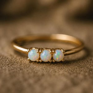 May include: A gold ring featuring three round, iridescent opals set in a horizontal row. The band is thin and polished, with each opal secured by small metal prongs. The stones display flashes of green, blue, and orange light against a white base.