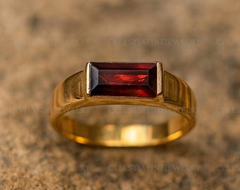 May include: A gold ring with a rectangular, deep red gemstone. The ring has a simple, classic design with a polished gold band and a bezel setting for the gemstone. The ring is displayed on a textured, neutral-toned surface.