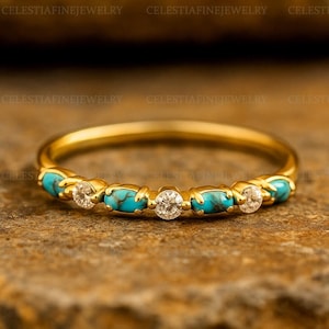 May include: A gold band ring with alternating turquoise and diamond-like stones. The ring is displayed on a textured, brown surface. The words "CELESTIAFINEJEWELRY" are visible in the background.