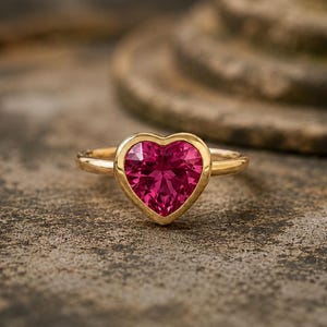 Op de afbeelding: Een gouden ring met een hartvormige, roze edelsteen. De edelsteen is gezet in een gouden bezel, met een effen gouden band. De ring wordt gepresenteerd op een getextureerd, grijs oppervlak.