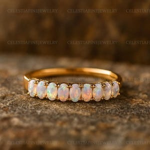 May include: A gold band ring featuring a row of oval-cut opals. The opals have a milky white base with flashes of color. The ring is set against a textured, brown background. The text "CELESTIAFINEJEWELRY" is visible.