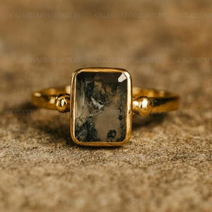 Puede incluir: Un anillo de oro con una piedra rectangular de ágata musgosa. La piedra tiene un patrón único de inclusiones verdes y grises. La banda del anillo es de oro pulido con detalles decorativos a cada lado de la piedra. El texto "CELESTIAFINEJEWELRY" es visible.