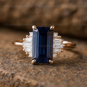 May include: A gold ring featuring a large, rectangular, deep blue gemstone as the centerpiece. The ring is accented with smaller, rectangular, and round white gemstones. The band is a warm gold color, and the text "CELESTIAFINEJEWELRY" is visible.