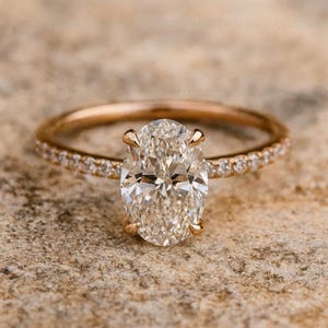 May include: A rose gold engagement ring with an oval-cut diamond center stone. The band is also rose gold and features small, round diamonds. The ring is set against a textured, neutral background.