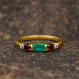 May include: A gold ring featuring a rectangular emerald-cut green gemstone flanked by two round red gemstones and two small white gemstones. The ring is set against a textured, dark brown background. The text "CELESTIAFINEJEWELRY" is visible.