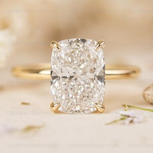 May include: A gold ring featuring a large, cushion-cut diamond. The diamond is clear and multifaceted, set with four prongs. The band is a thin, polished gold. The ring is displayed on a neutral background.