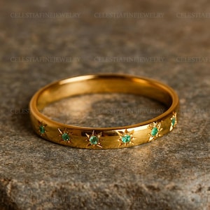 Puede incluir: Un anillo de banda dorado con un patrón repetitivo de estrellas, cada una con una pequeña gema verde redonda. El anillo se muestra sobre una superficie gris texturizada. El nombre de la marca "CELESTIAFINEJEWELRY" está impreso alrededor del anillo.