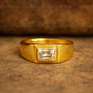 Op de afbeelding: Een gouden ring met een rechthoekige, heldere edelsteen in het midden. De ring heeft een dikke band en een vierkante kop. De woorden "CELESTIAFINEJEWELRY" zijn rond de ring gedrukt.