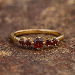 Peut inclure: Une bague en or ornée de cinq pierres précieuses rouges. Les pierres sont serties sur un fin anneau. La pierre centrale est légèrement plus grande que les autres. La bague est présentée sur une surface brune texturée. Le texte "CELESTIAFINEJEWELRY" est visible.