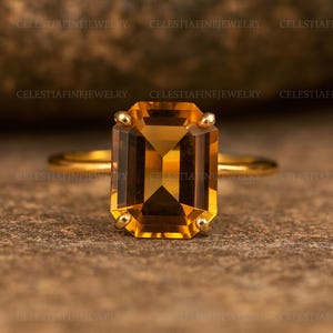 Peut inclure: Une bague en or ornée d'une grande pierre précieuse rectangulaire de couleur ambre. La pierre a une taille à facettes et la bague a de petits accents dorés à chaque coin. La bague est posée sur un fond neutre.