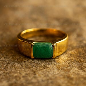 May include: A gold-toned ring with a rectangular, emerald-green stone. The ring has a thick band and a classic, elegant design. The stone is set flush within the ring's frame, creating a smooth, polished look. The ring is displayed on a textured, neutral-toned surface.