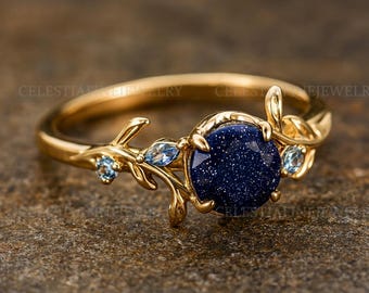 Könnte beinhalten: Ein goldfarbener Ring mit einem großen, runden, dunkelblauen Edelstein mit funkelnden Einschlüssen. Der Ring hat ein florales Design mit kleinen blauen Akzentsteinen und blattartigen Details. Der Ring ist vor einem neutralen Hintergrund platziert.