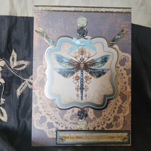 May include: A greeting card with a dragonfly design. The card features a light blue and gold frame with a dragonfly illustration. The text "MAY ALL YOUR Birthday Wishes COME TRUE" is at the bottom. The card has a lace-like pattern.