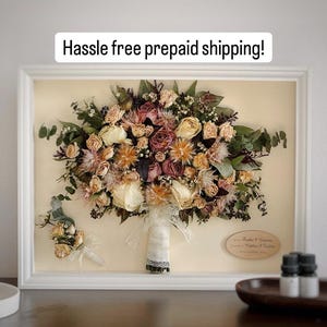 May include: A framed bridal bouquet and boutonniere, preserved and displayed. The bouquet features dried roses and other flowers in cream, peach, and burgundy, with green foliage. The white frame surrounds the arrangement. Text at the top reads "Hassle free prepaid shipping!"