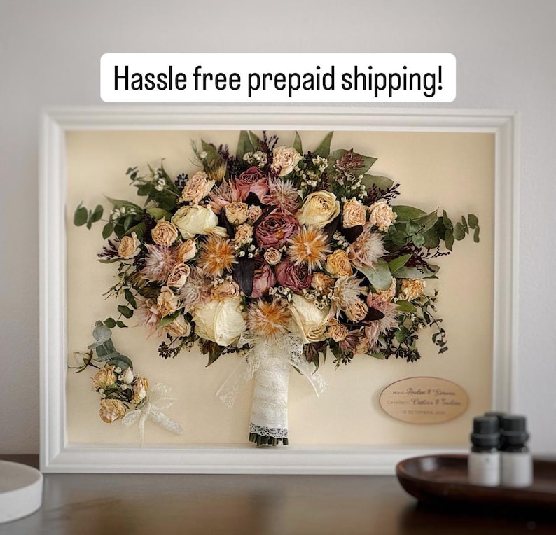 May include: A framed bridal bouquet and boutonniere, preserved and displayed. The bouquet features dried roses and other flowers in cream, peach, and burgundy, with green foliage. The white frame surrounds the arrangement. Text at the top reads "Hassle free prepaid shipping!"