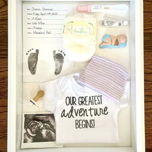 May include: A framed baby keepsake shadow box with a white frame. The box contains a baby's footprint, a diaper, a onesie with the text "OUR GREATEST ADVENTURE BEGINS!", a baby hat, and a sonogram. The baby's length was 19 inches.