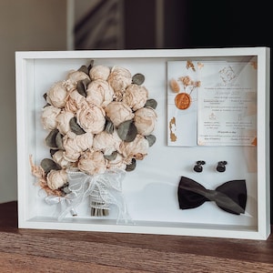 May include: A white framed wedding memento. It contains a bouquet of cream-coloured flowers, a wedding invitation, a black bow tie, and cufflinks. The bouquet is tied with a white ribbon. The invitation has gold accents and text.