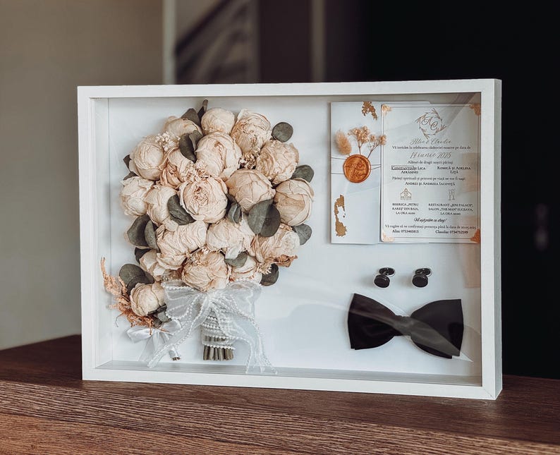 May include: A white framed wedding memento. It contains a bouquet of cream-coloured flowers, a wedding invitation, a black bow tie, and cufflinks. The bouquet is tied with a white ribbon. The invitation has gold accents and text.