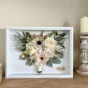 May include: A framed floral arrangement featuring a bouquet of preserved flowers in shades of white, cream, and pink. The bouquet is accented with green foliage and a white ribbon. A small wooden plaque is attached to the frame.