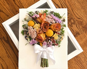 May include: A framed wall art piece featuring a pressed bouquet of dried flowers including orange roses, yellow billy balls, and purple statice. The stems are tied with a white satin ribbon bow, all mounted on a white rectangular canvas.