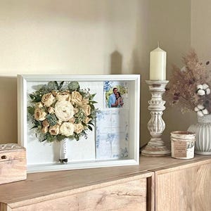 May include: A framed wedding bouquet with a photo and invitation. The bouquet features cream-colored roses and greenery. A wooden box, candle, and vase with dried flowers are on a wooden cabinet.