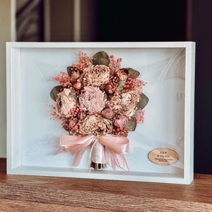 May include: A framed display of dried flowers, including pink and beige roses, with green foliage and red berries. The bouquet is tied with a pale pink ribbon and set in a white shadow box frame, ideal for home decor.