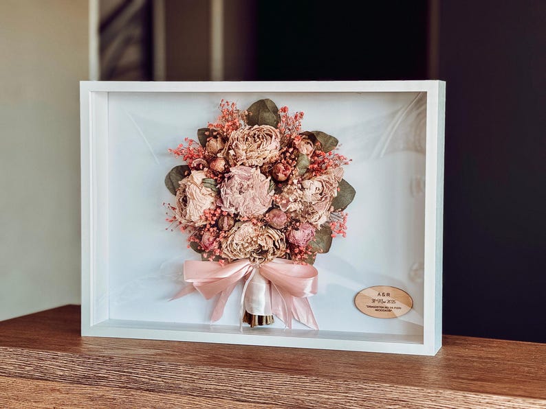 May include: A framed display of dried flowers, including pink and beige roses, with green foliage and red berries. The bouquet is tied with a pale pink ribbon and set in a white shadow box frame, ideal for home decor.