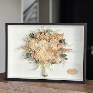 May include: A framed display of a dried flower bouquet in cream, beige, and green tones. The bouquet is tied with a pale green ribbon and set against a white backdrop within a black frame. A small wooden plaque with the date 29.05.2025 is visible.