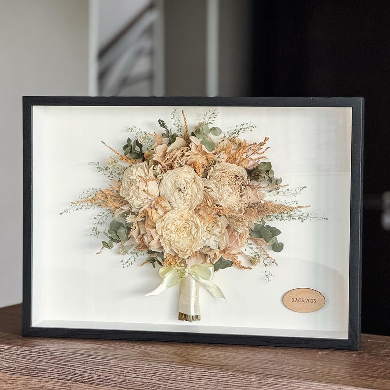 May include: A framed display of a dried flower bouquet in cream, beige, and green tones. The bouquet is tied with a pale green ribbon and set against a white backdrop within a black frame. A small wooden plaque with the date 29.05.2025 is visible.