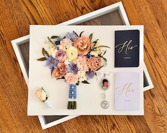 May include: A framed wedding keepsake display featuring a dried floral bouquet with peach, lavender, and white flowers, a single peach boutonniere, a navy blue 'His Vows' booklet, a light purple 'Her Vows' booklet, and a silver photo charm pendant.
