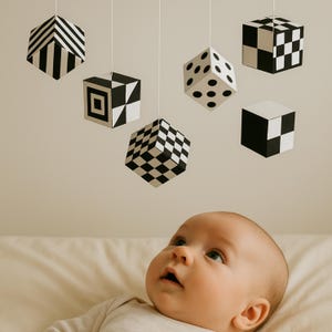 Könnte beinhalten: Ein Baby blickt auf ein Mobile mit sechs geometrischen Würfeln in Schwarz und Weiß. Die Würfel weisen verschiedene Muster auf, darunter Streifen, Karos und Punkte. Das Mobile hängt an weißen Schnüren vor einem neutralen Hintergrund.
