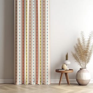 May include: Cream-colored curtain with vertical stripes in various colors, including red, green, and blue. The stripes are punctuated by small black rectangles. The curtain is in front of a white wall, with a small wooden table and decorative vase.
