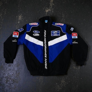 Op de afbeelding: Zwarte, blauwe en witte racejas met sponsorlogo's, waaronder "Castrol" en "Ford". De jas heeft een volledige ritssluiting, een opstaande kraag en "Ford Performance Racing" verticaal gedrukt.