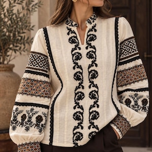 May include: An off-white long-sleeve blouse featuring intricate black floral embroidery along the center placket and sleeves. The garment includes black lace trim accents, a mandarin collar with a small V-neck opening, and gathered cuffs.