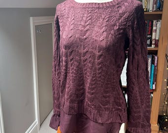 May include: A long-sleeve jumper in a deep burgundy hue featuring a vertical cable knit pattern on the front and sleeves. The garment has a round neckline and a layered hemline with a smooth, solid-coloured fabric panel extending below.