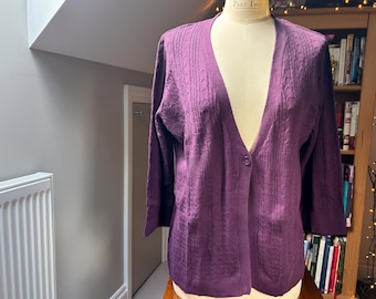 May include: A deep purple V-neck cardigan featuring a single button closure at the centre. The knit fabric displays vertical cable knit patterns and ribbed detailing along the front panels and sleeves, providing a textured appearance.