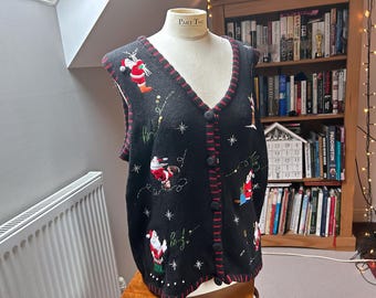 May include: A black V-neck sweater vest featuring embroidered Santa Claus figures, white star accents, and the word "ho" stitched in green. The vest has a button-front closure with red and black striped trim along the edges and armholes.