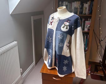 May include: A zip-up knit cardigan featuring a colour-blocked design in cream and various shades of blue. The garment is decorated with embroidered snowman figures wearing scarves and hats, interspersed with white stitched snowflake patterns.