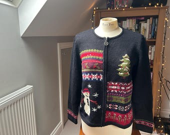 May include: A black knit zip-up cardigan featuring a festive holiday design. The front displays a snowman, a decorated Christmas tree, and colourful horizontal bands with snowflake and geometric patterns. It has a round neckline and long sleeves.
