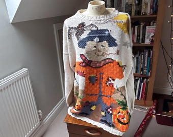 May include: A cream-coloured long-sleeve knit jumper featuring a large intarsia design of a scarecrow wearing a blue hat and orange shirt, with a yellow sun, a black crow, and a jack-o'-lantern pumpkin at the bottom hem.