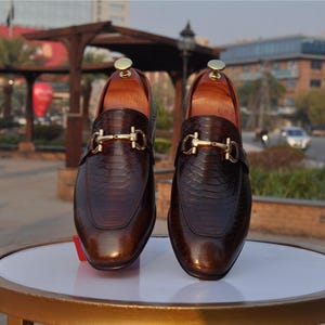 May include: A pair of brown leather loafers with a snakeskin texture. The shoes feature a gold-coloured buckle and are displayed on a white surface. The shoes are designed for formal wear.